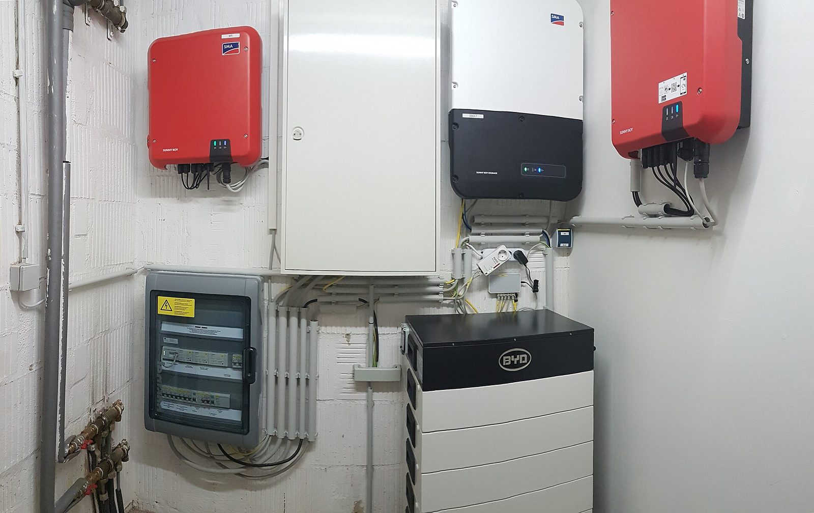 Technische ruimte van een woning met twee rode omvormers, een batterijomvormer, een batterijtoren op de vloer en een zekeringkast