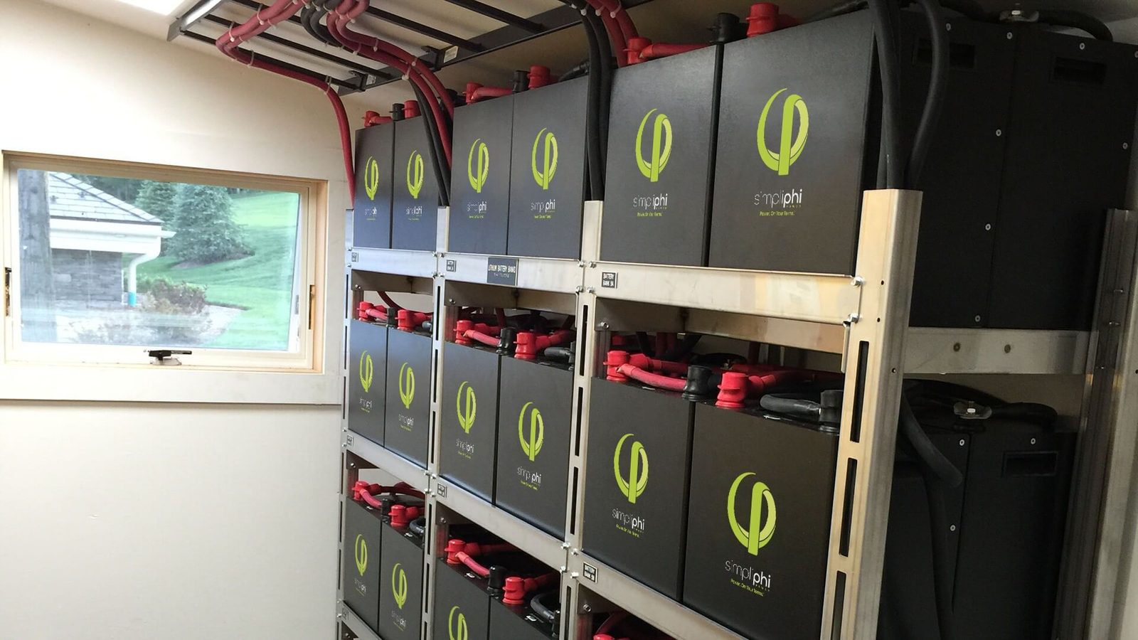 Rek met een reeks zwarte batterijmodules en rode kabels in een aparte technische ruimte met een raam