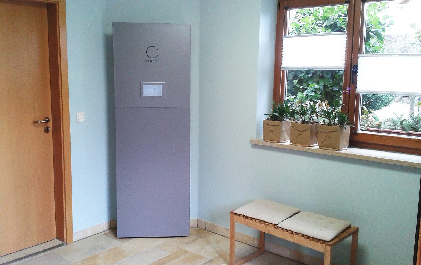 Grijze staande thuisbatterij met aanraakscherm in een lichtblauwe kamer, naast een deur, een raam met planten en een houten bankje